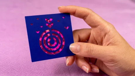A hand holding a navy business card die-cut with a bullseye pattern and small hearts, revealing bright pink foil underneath