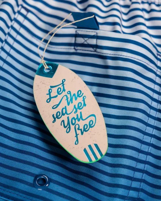 A cream, surfboard-shaped hang tag with teal foil script reading Let the sea set you free, hanging by a cord from blue-and-white striped swim-shorts fabric.