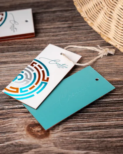 Two foil hang tags with a geometric maze logo and 'Creative' script wordmark, cream and teal, hanging on a wood surface.