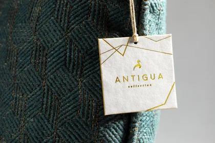 A white hang tag with gold foil geometric line-art and Antigua Collection lettering, hanging by a cord from a teal woven-texture fabric.