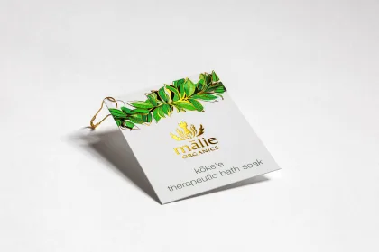 A white, gold-foil hang tag for Mālie Organics with a green and gold foil laurel-leaf branch across the top, clipped on with a small gold pin.