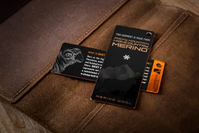 Three foil hang tags on a leather-like brown surface: a black tag with a ram graphic, a black tag reading 'Rocky Mountain Highcountry Merino,' and an orange tag with a snowflake logo.