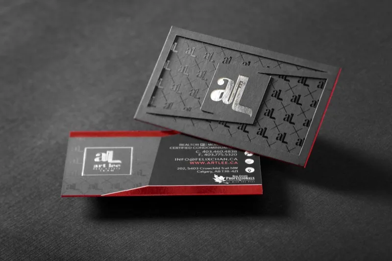 A layered dark gray real-estate business card with a silver foil monogram and red colored edges, standing upright beside the closed card.