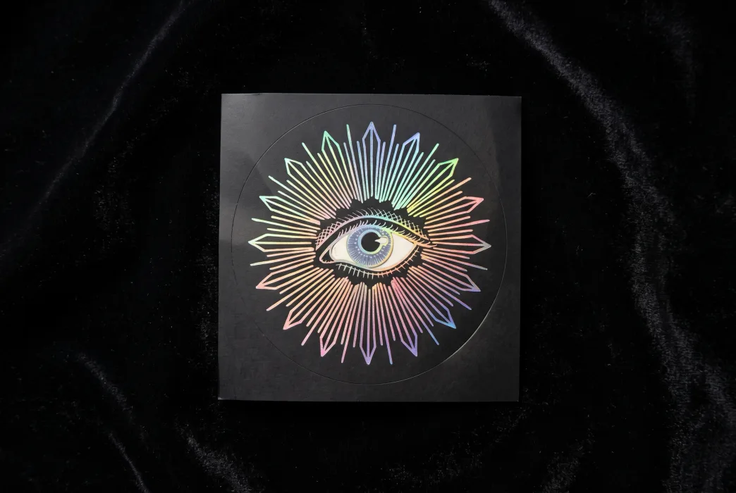 A round foil sticker with a rainbow holographic sunburst and eye design, the metallic pattern shifting color against black velvet.